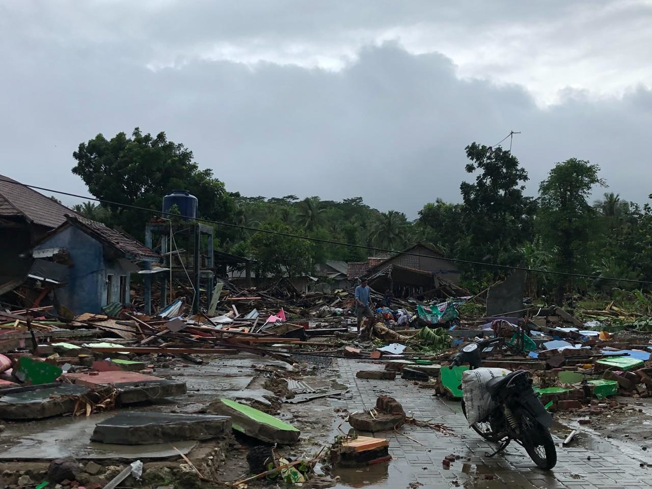 A man stands amid debris from destroyed houses scattered across the ground, with a motorbike visible in the corner of the frame. Gray skies and clouds loom overhead.