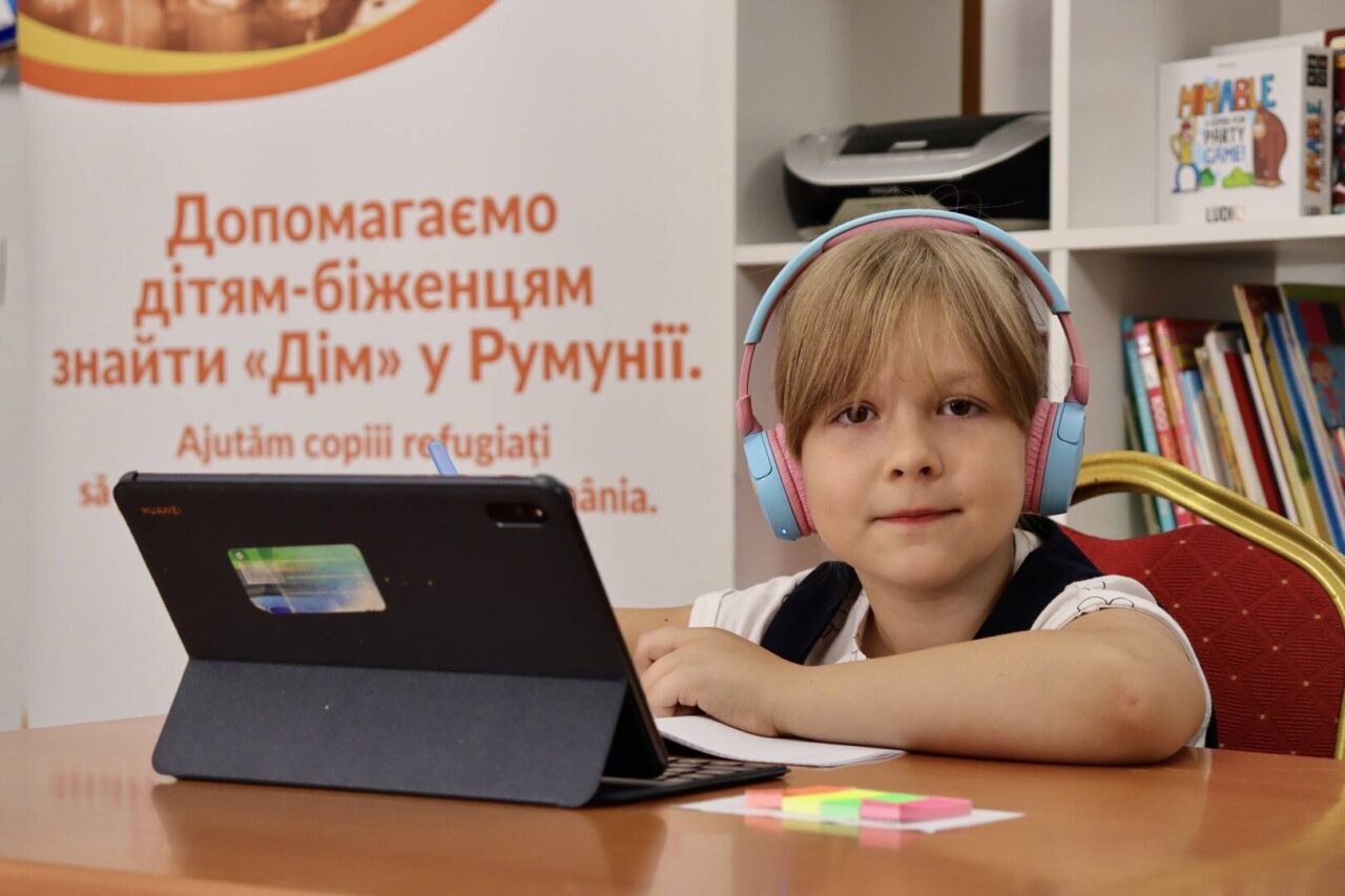 A young blonde girl faces the camera, seated in a learning space. She&rsquo;s wearing headphones, and an open laptop sits before her.