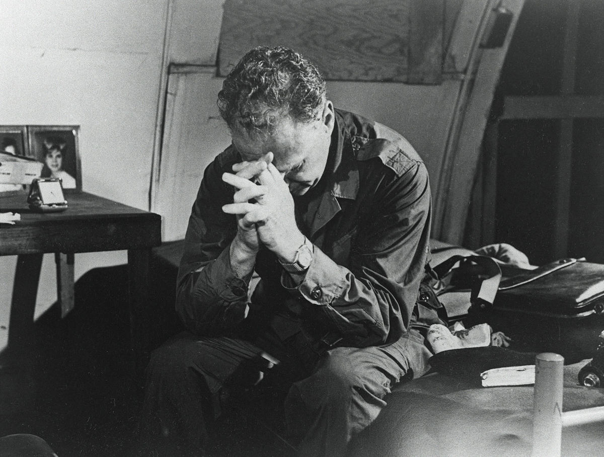A man sits on a bed with his head bowed and hands clasped in prayer beside a table with family photos.