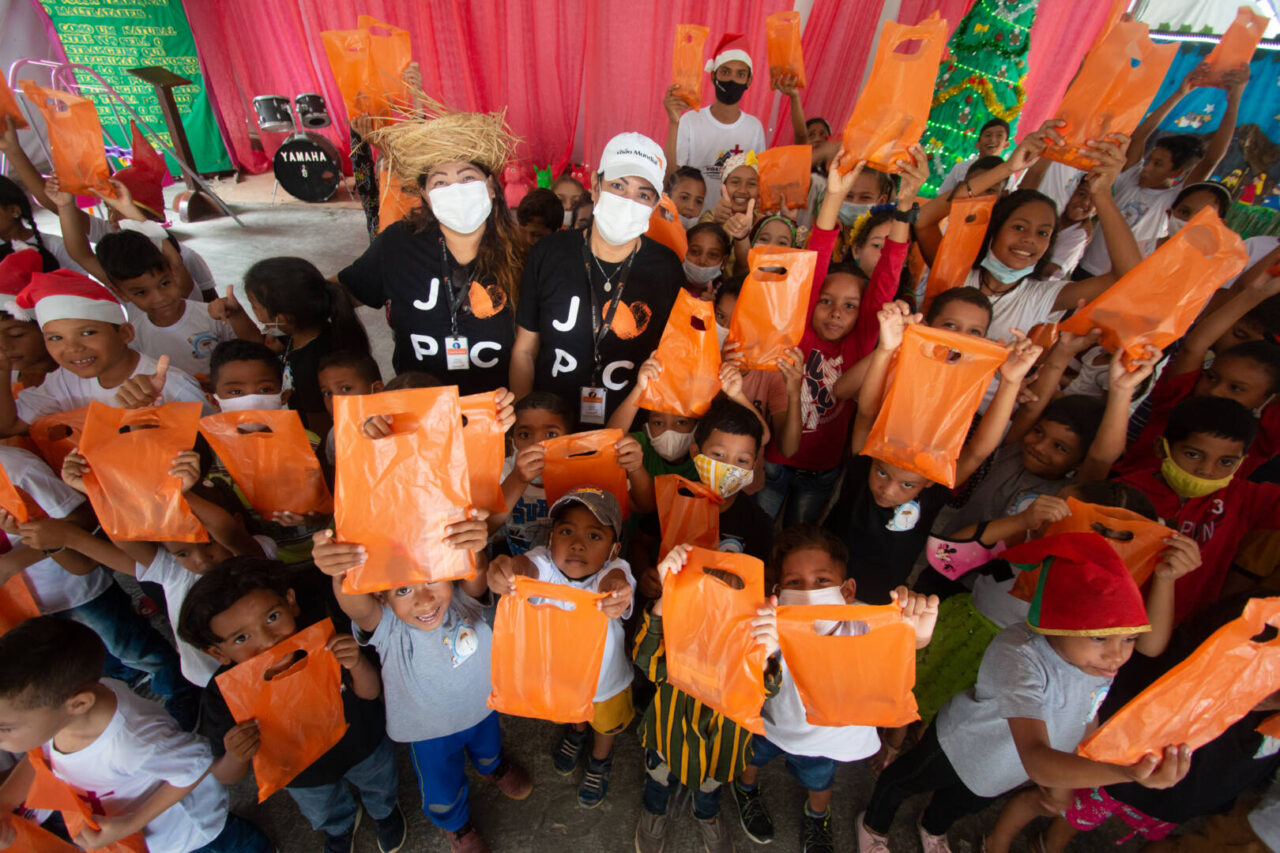 A large group of young children hold up orange plastic bags to the camera during a Christmas party in Brazil.