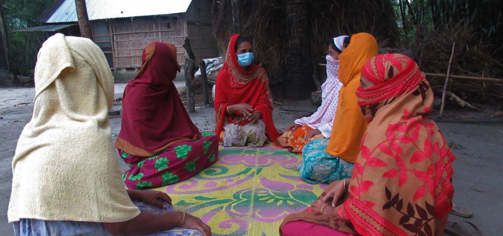 Community meeting Six Bangladeshi women in colorful attire sit on a colored mat outside.