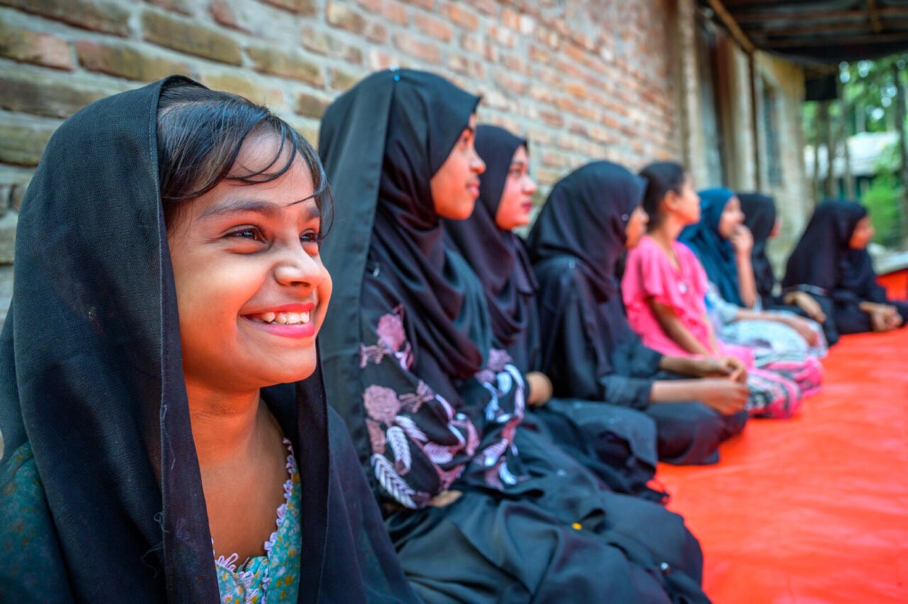 A group of girls of varying ages, most wearing headscarves, look and listen to a person speak, who is not visible. They sit against a brick wall.