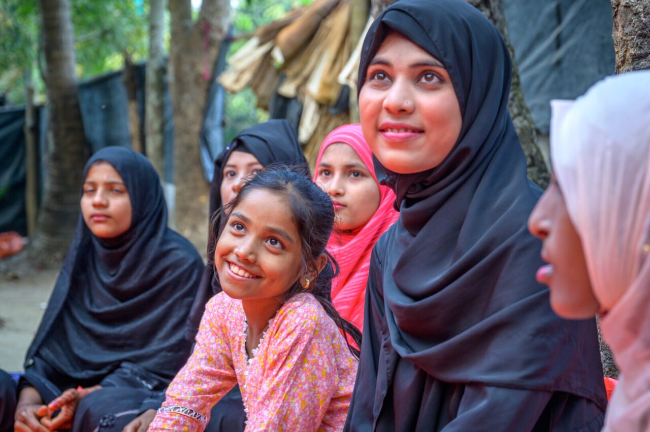 A group of girls of varying ages, most wearing headscarves, look up and listen to a person off camera.