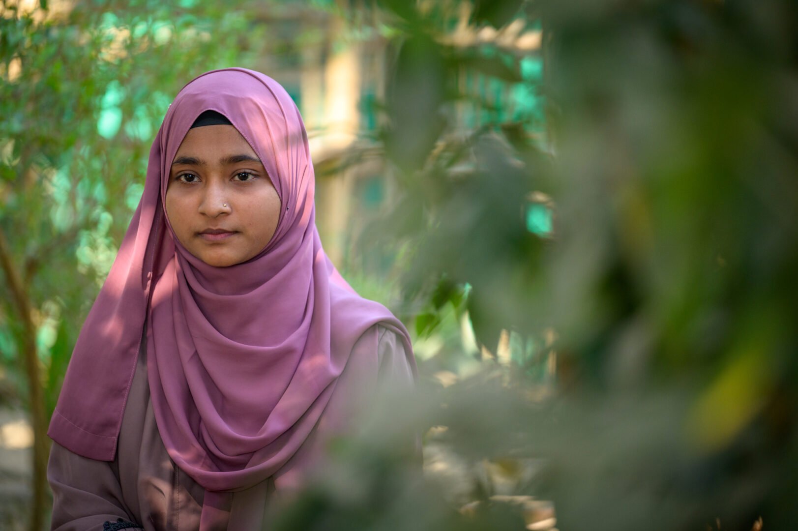 A young woman in a pink headscarf stands in a dirt walkway bordered by bamboo fences.
