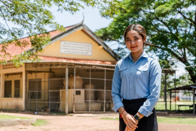 Phanet wears a light blue shirt stands smiling outside a school building with a red-tiled roof and yellow walls.