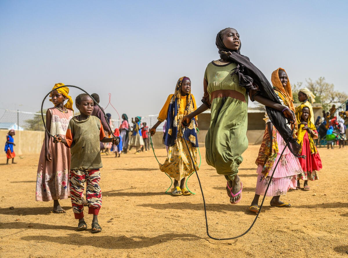A girl in a headscarf and green dress jumps rope, mid-air, surrounded by other children jumping rope.
