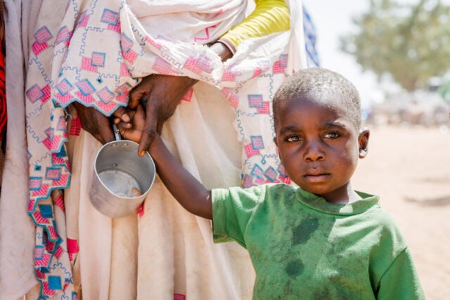 A young boy in a green T-shirt looks at the camera while holding a woman's hand. An empty tin cup dangles from her fingers.