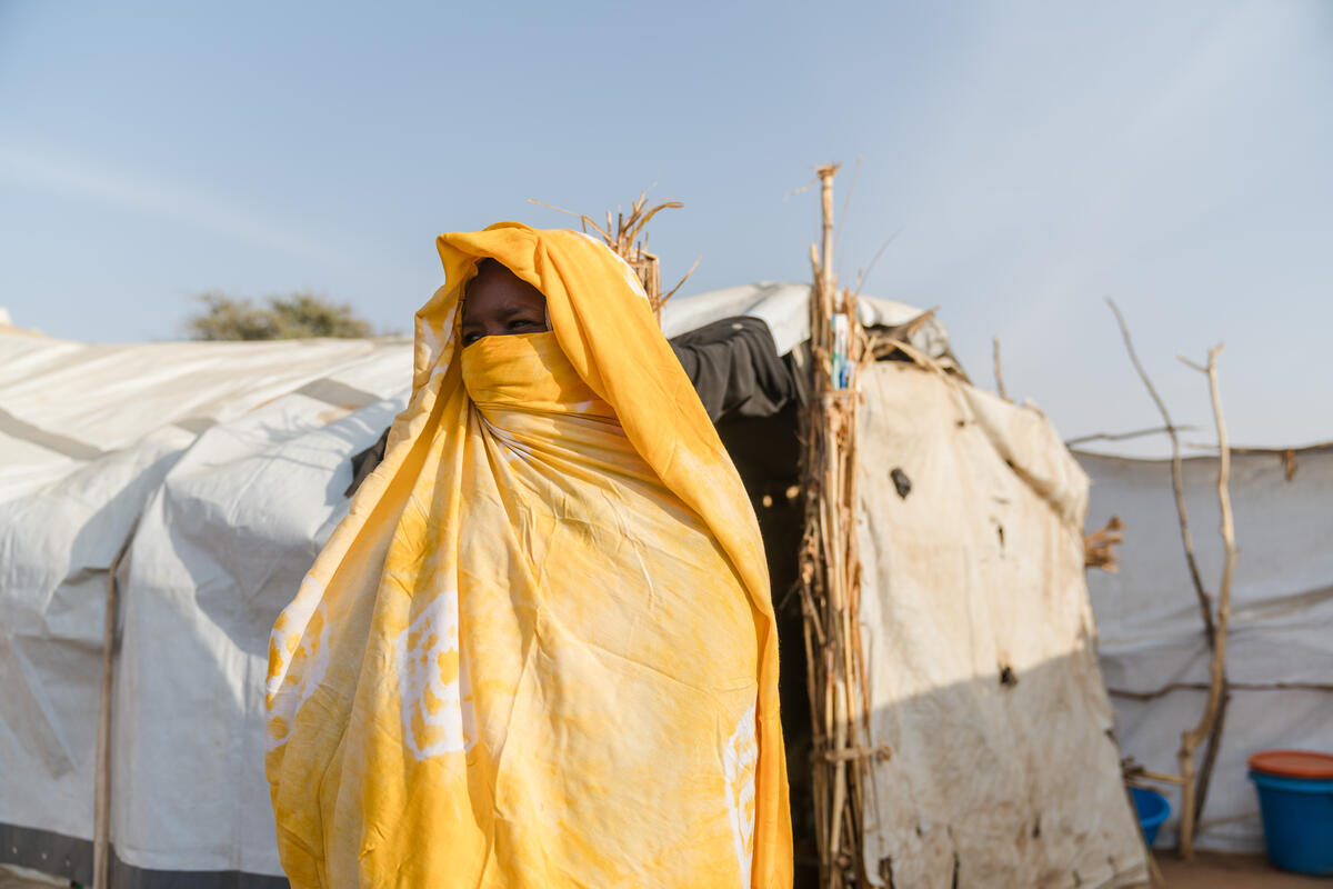 A woman wrapped in a yellow shawl stands in front of makeshift shelters in a displacement camp under a clear sky.