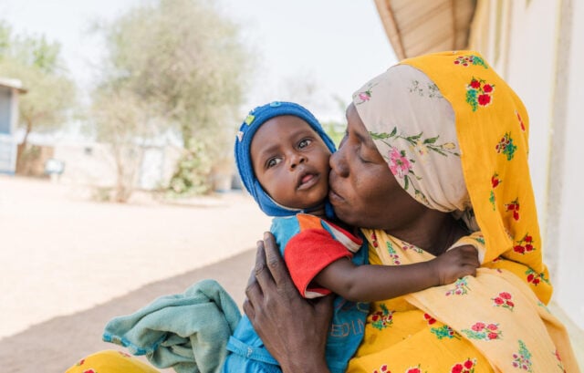 : A woman kisses her baby on the cheek while holding him close outside a building. A tree is in the background, and the ground is very dry.