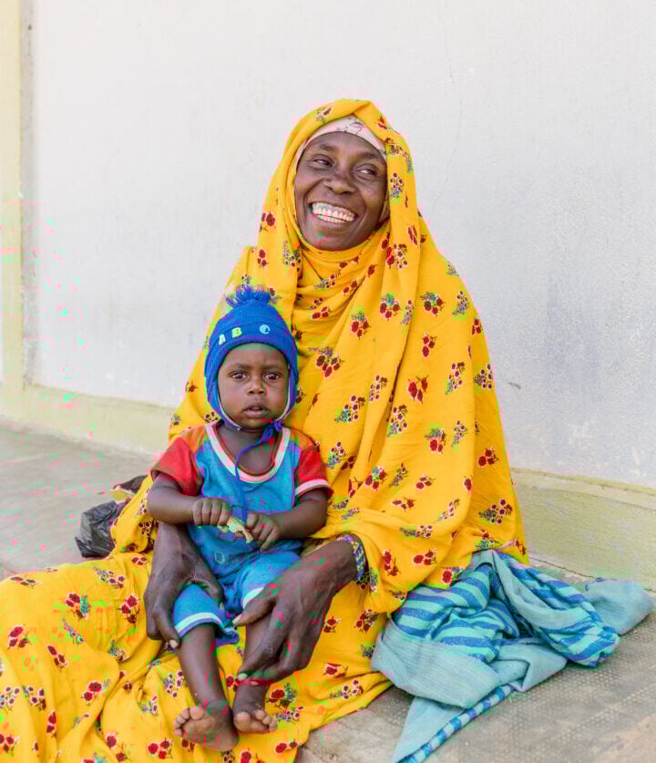 A smiling woman in a bright yellow head scarf holds her young child, who wears a blue hat, as they sit against a wall.