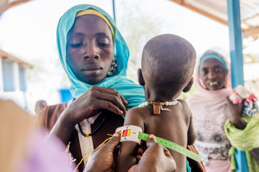 A woman holds her young child while a health worker measures the child’s arm for malnutrition. Another woman looks on in the background.