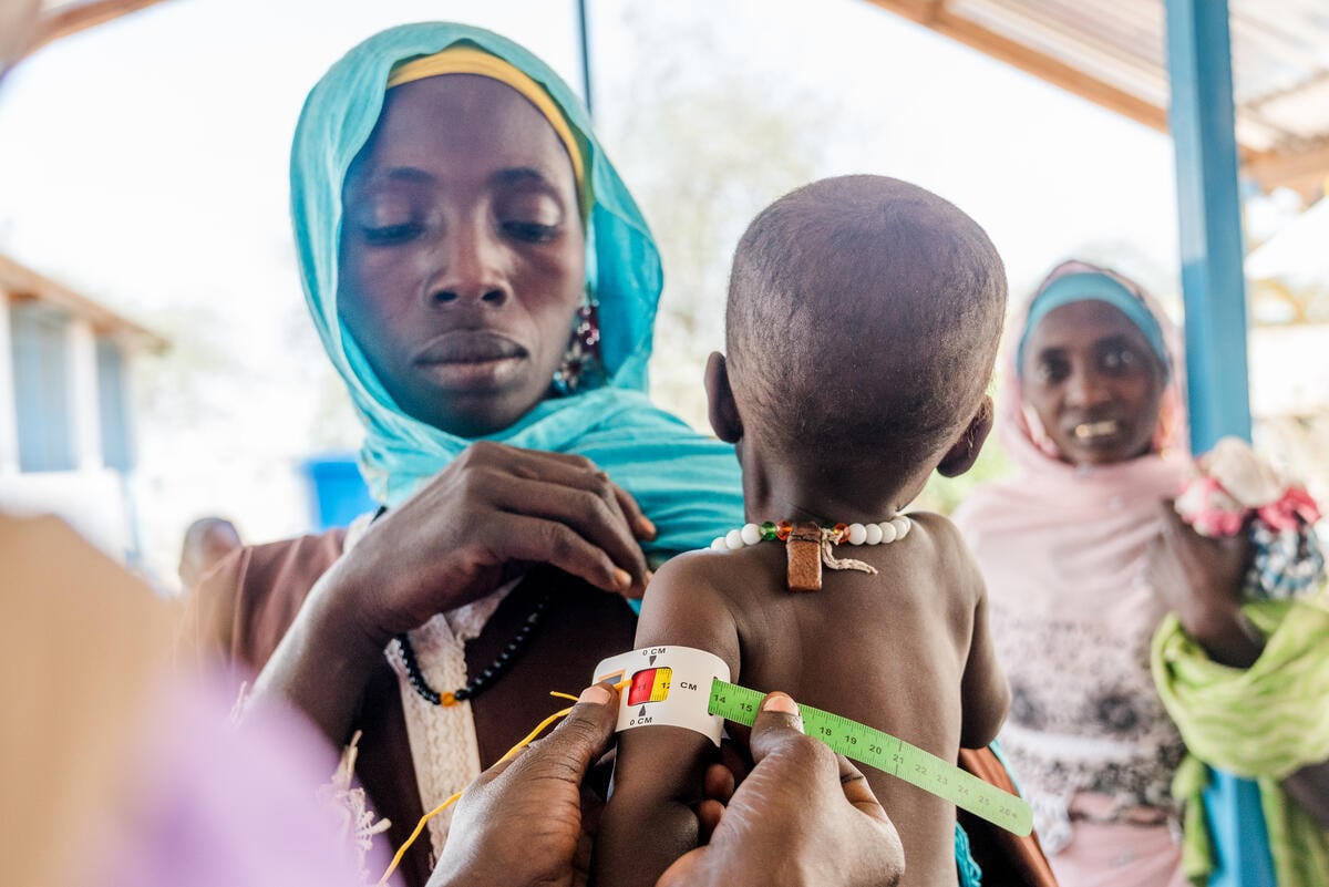 A woman holds her young child while a health worker measures the child’s arm for malnutrition. Another woman looks on in the background.