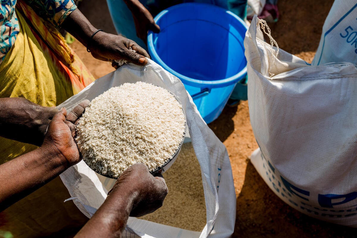 Hands holding a bowl of rice, with an empty blue plastic bin and an opened sack with grain resting nearby, shows a moment of food distribution in Chad.
