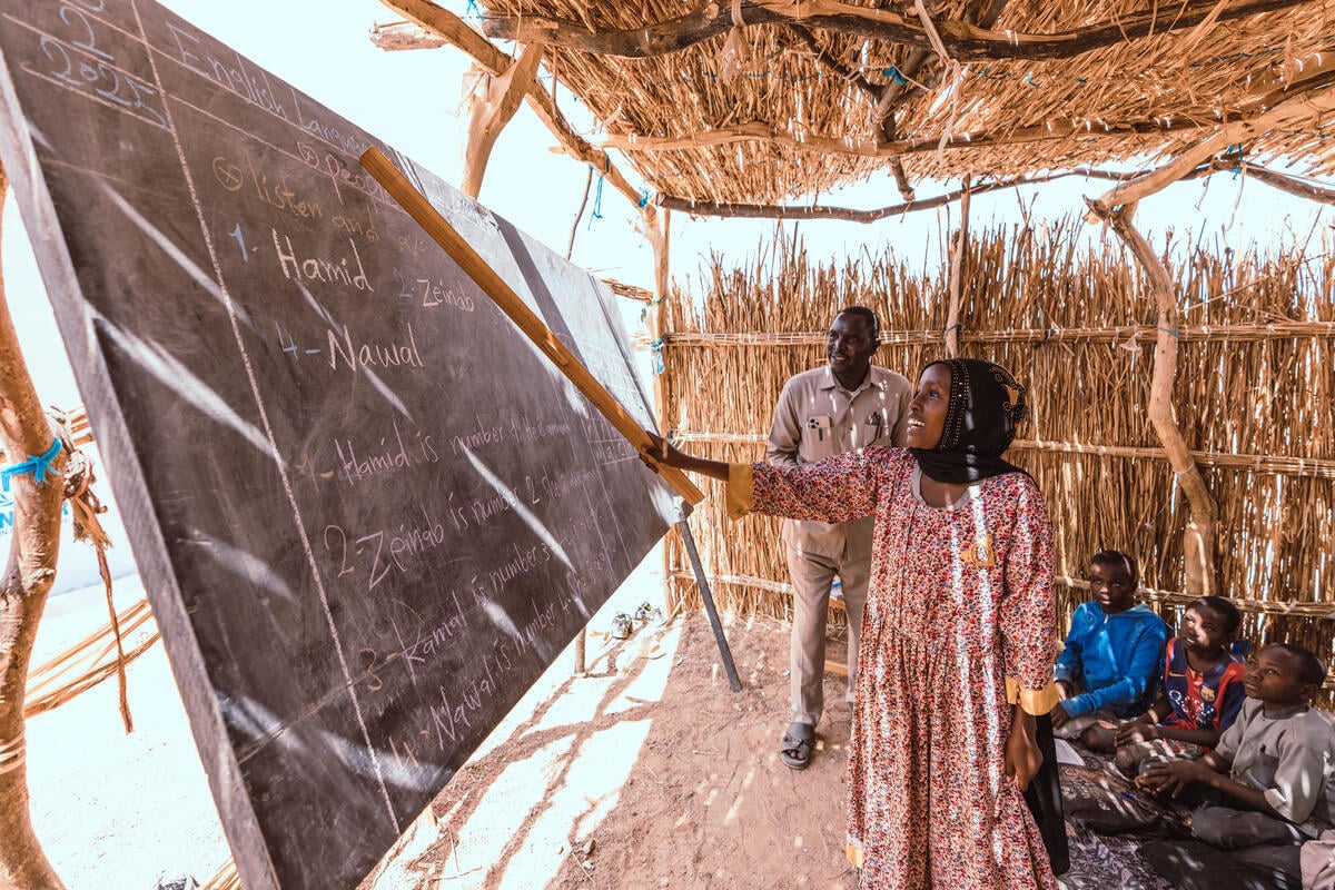 A young girl uses a wooden ruler to point at a chalkboard with English text in an outdoor classroom as children sit on the ground behind.
