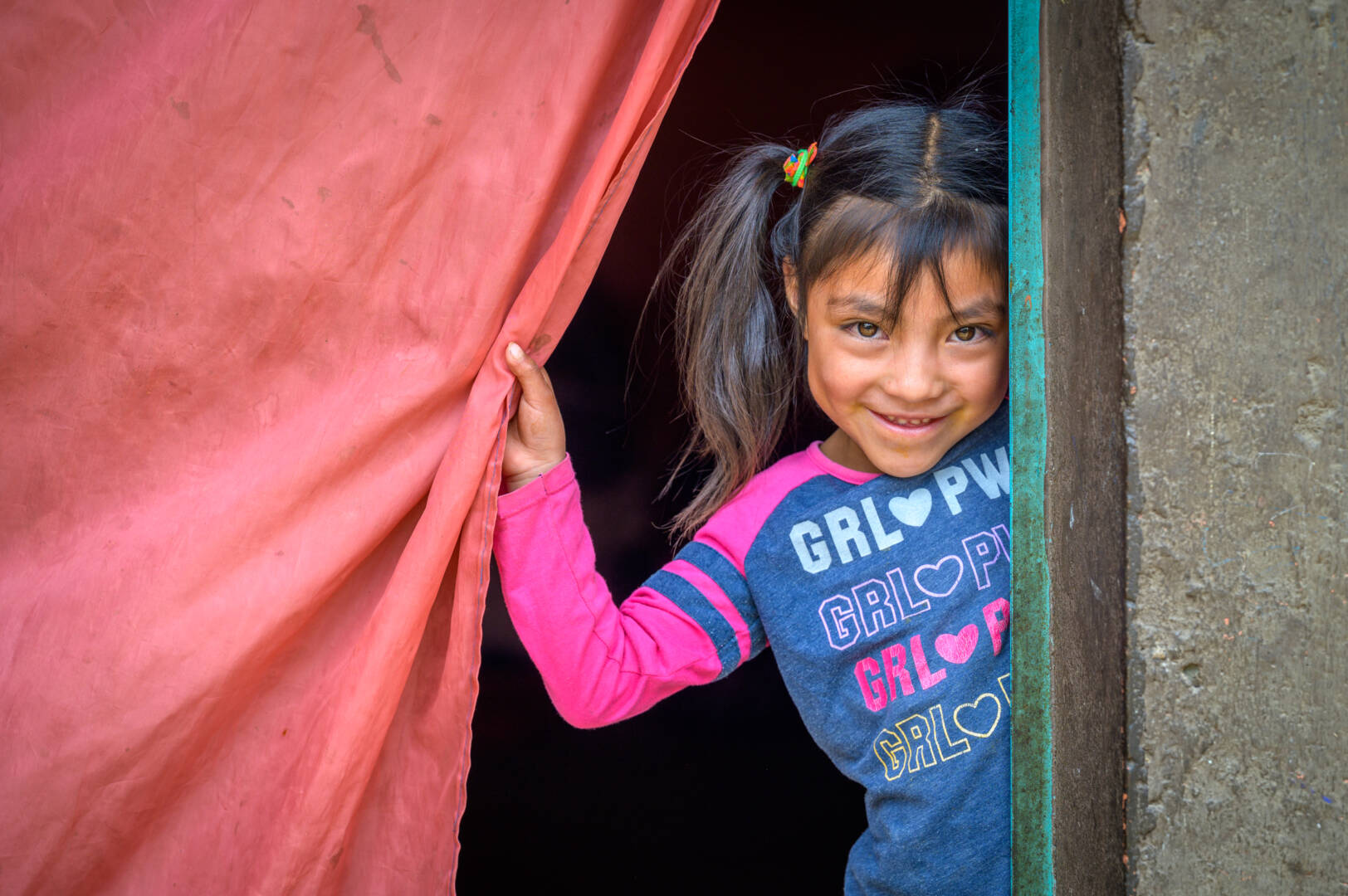 A girl in pigtails smiles from a doorway while holding a red curtain aside.