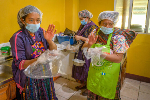 Women wearing aprons and hairnets stand in a brightly colored kitchen and pound chocolate paste in bags with their hands.