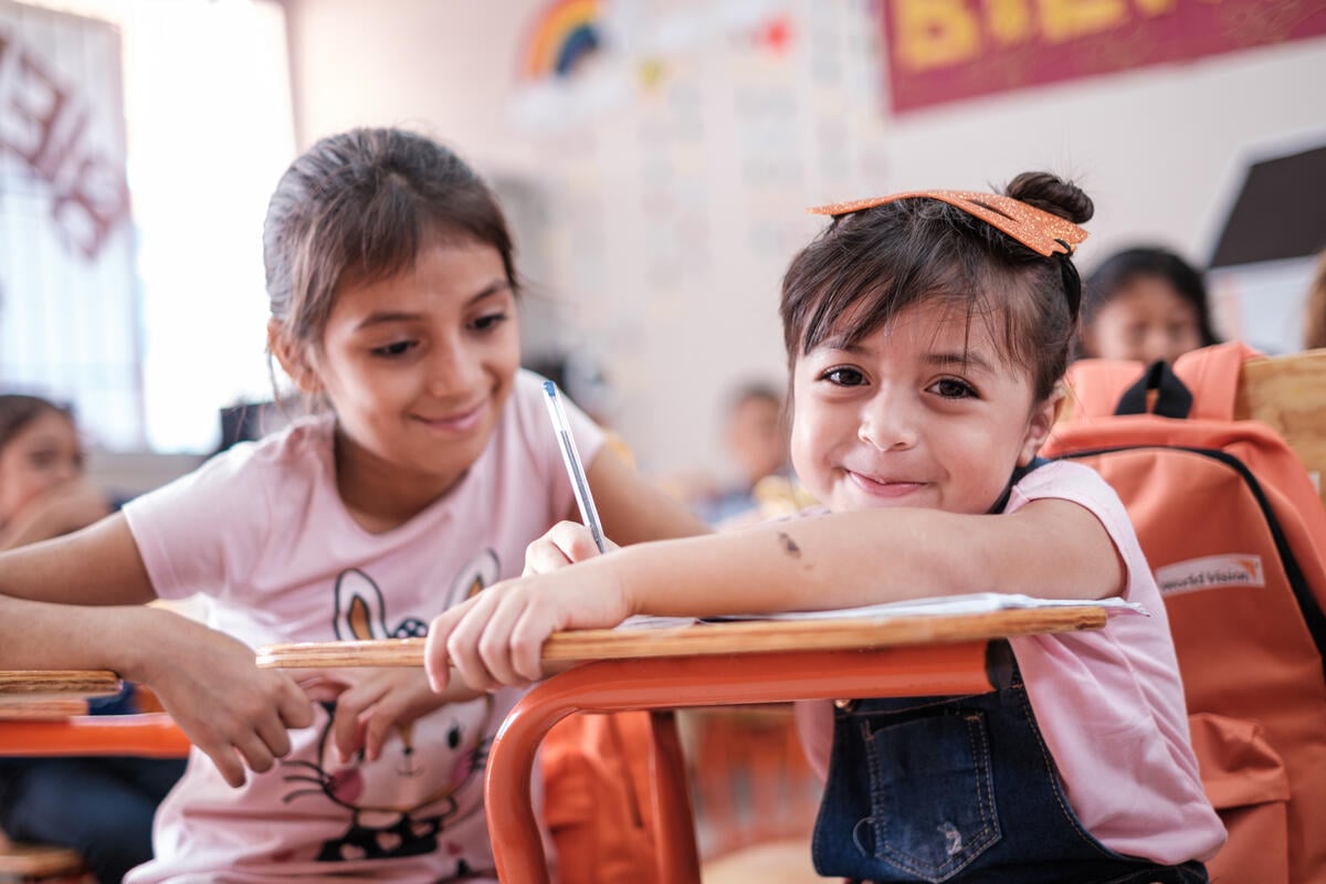 Two young girls dressed in pink smile brightly in a classroom. One girl looks into the camera, while the other gazes at her classmate.