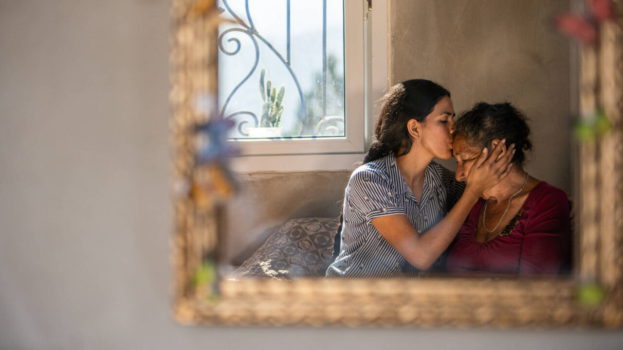 A mirror captures a reflection of a young woman holding an elderly woman&rsquo;s face in her hands while she gently kisses the woman&rsquo;s forehead.