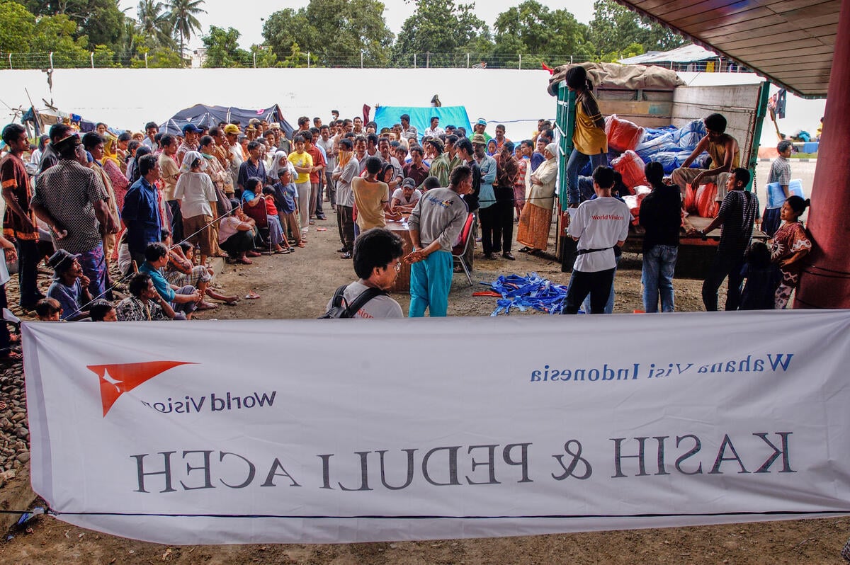 A crowd gathers for an aid distribution in Indonesia, where a World Vision banner, visible from the backside, reads "Kasih & Peduli Aceh," highlighting humanitarian efforts.