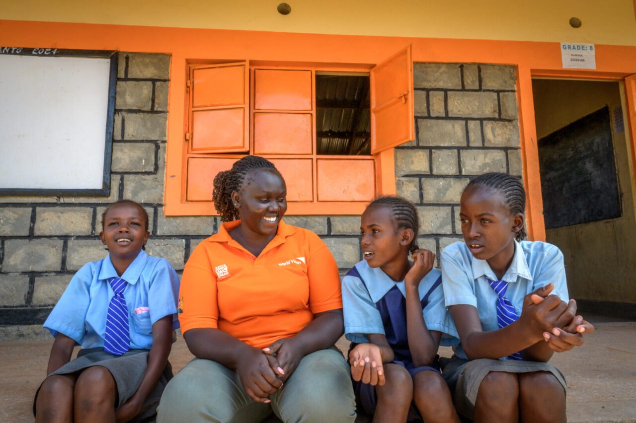 A female World Vision staff member sits on steps outside a classroom talking with three girls wearing school uniforms.