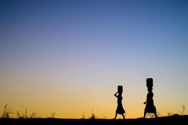 Woman and girl silhouetted against the sunset carry buckets on their heads.