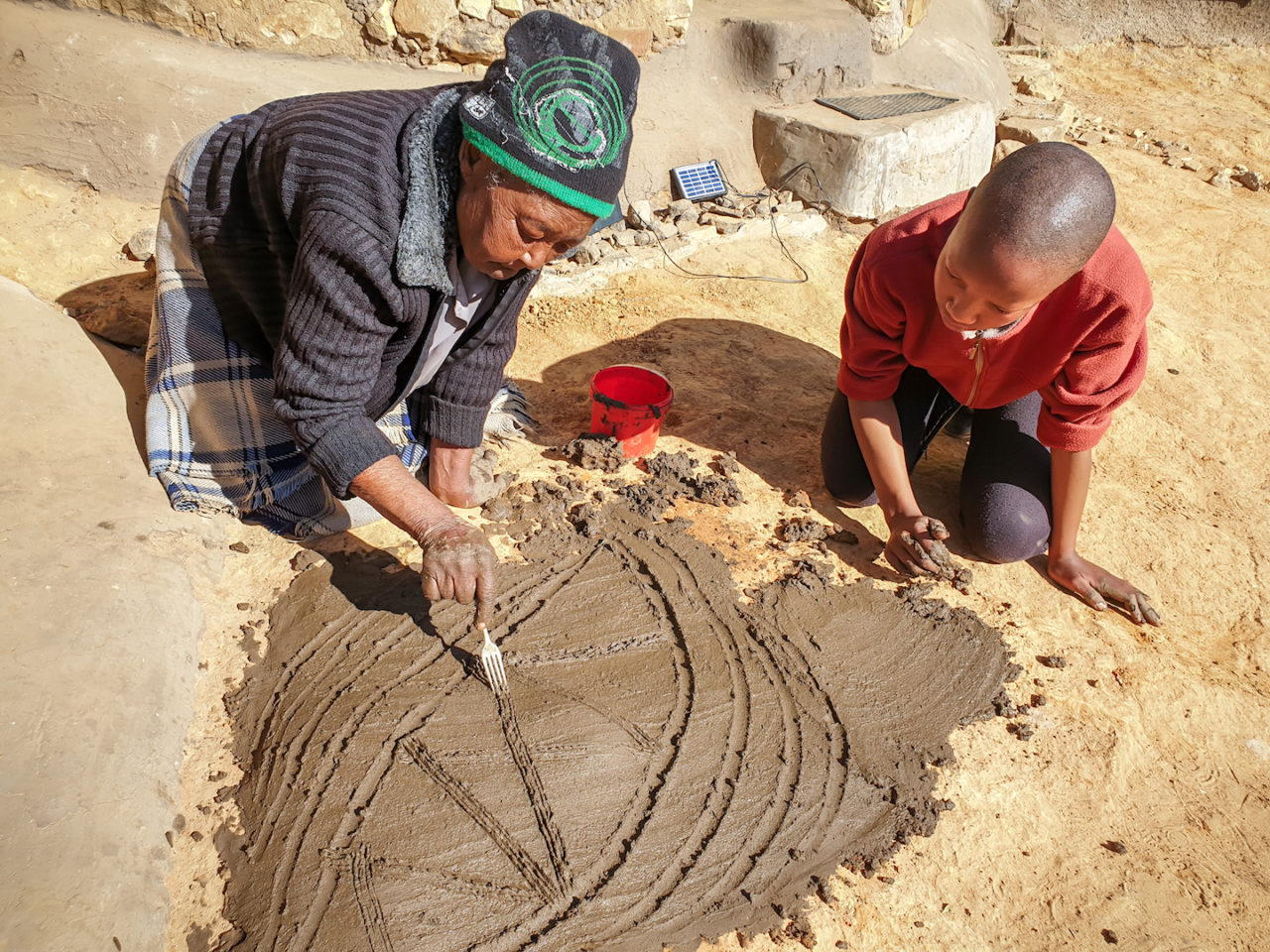 Woman and child decorate a patch of mud outside the entrance of their house. 