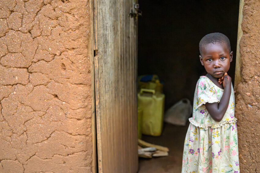 A young girl wearing a yellow floral dress stands in the doorway of a mud home in a rural area.