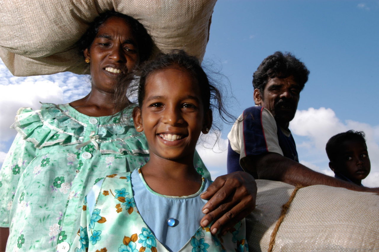 A smiling girl looks at the camera while a woman stands behind her, resting her hand on the child's shoulder. 