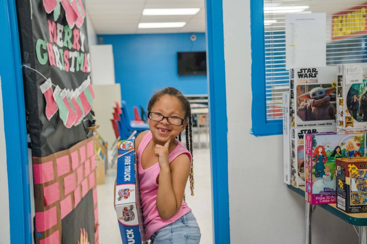 Standing in the doorway of a storage room full of toys and games, a little girl in glasses and a pink top smiles happily at the camera, clutching the boxed game she chose from the gift items.