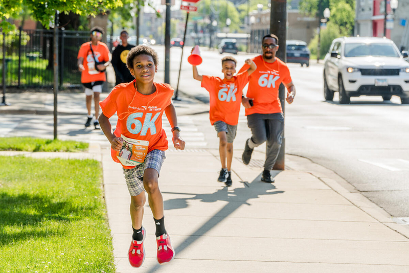 Two children and an adult run on a sidewalk in a city. They look happy, and one has his arms raised cheering.