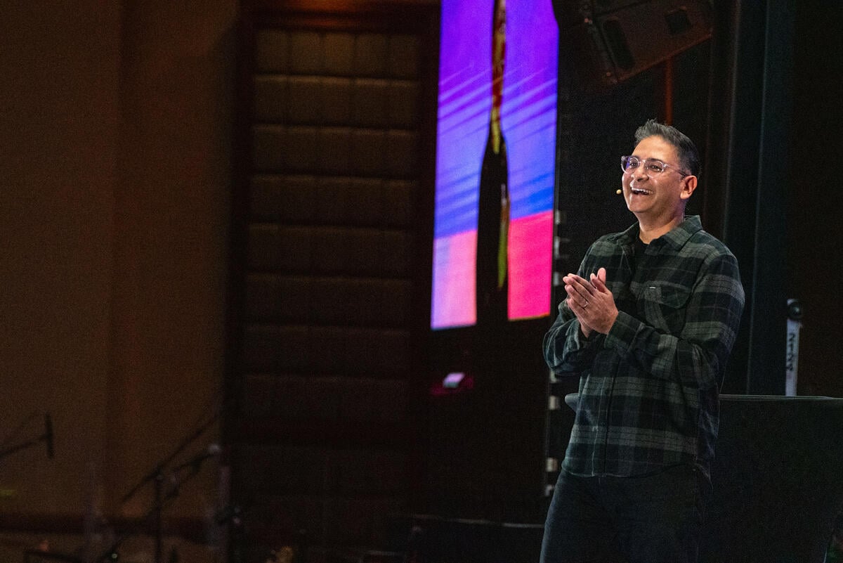 A man wearing glasses and a microphone headset smiles and claps while speaking on stage beside a large screen.