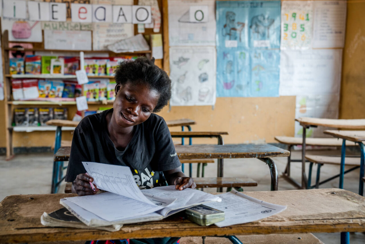 A young woman looks through a workbook while sitting at a desk in a classroom.