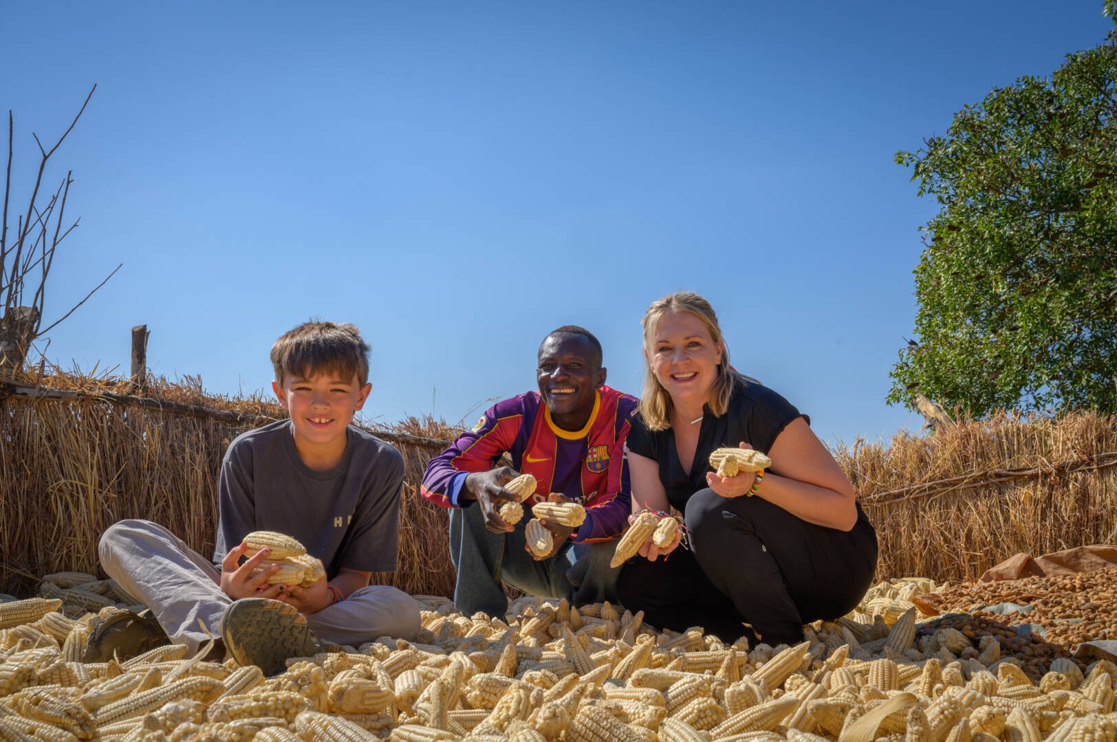 Melissa Joan Hart, a man, and a boy crouch among cobs of corn. They each hold several ears of corn in their hands.