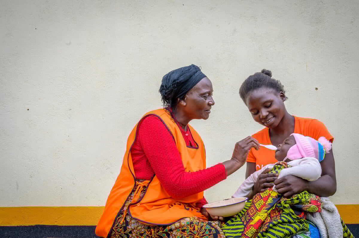 A woman in an orange vest spoonfeeds a baby in a pink knit cap, who is sitting on a smiling woman&rsquo;s lap.