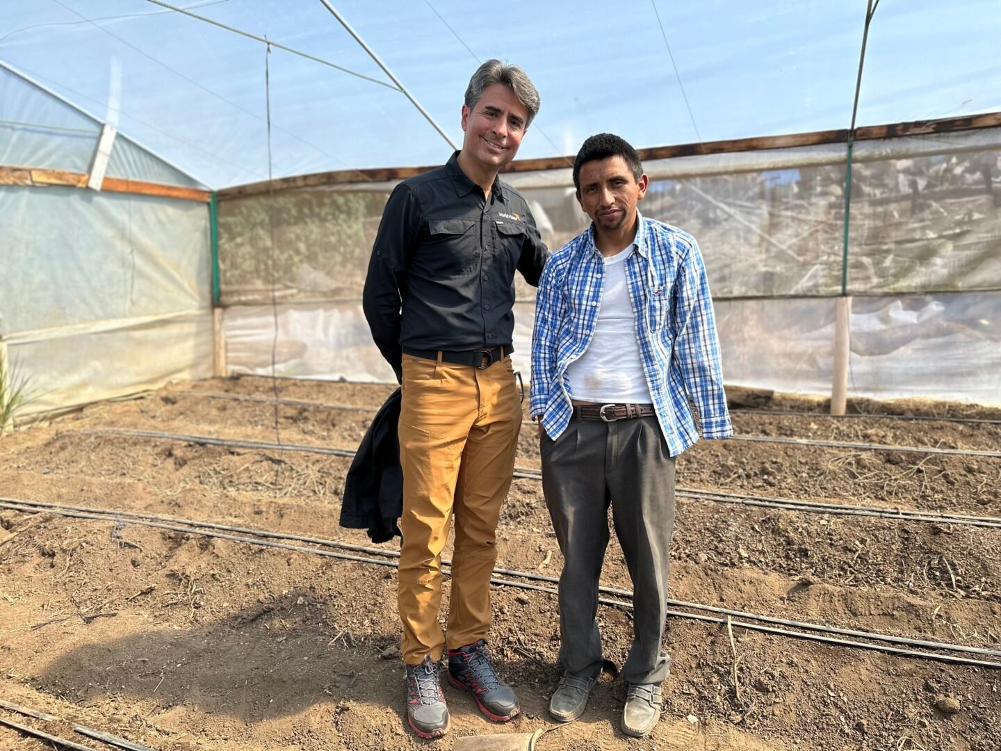 Two men stand side by side inside a greenhouse, posing on dry soil rows beneath a structure of plastic sheeting and wood.