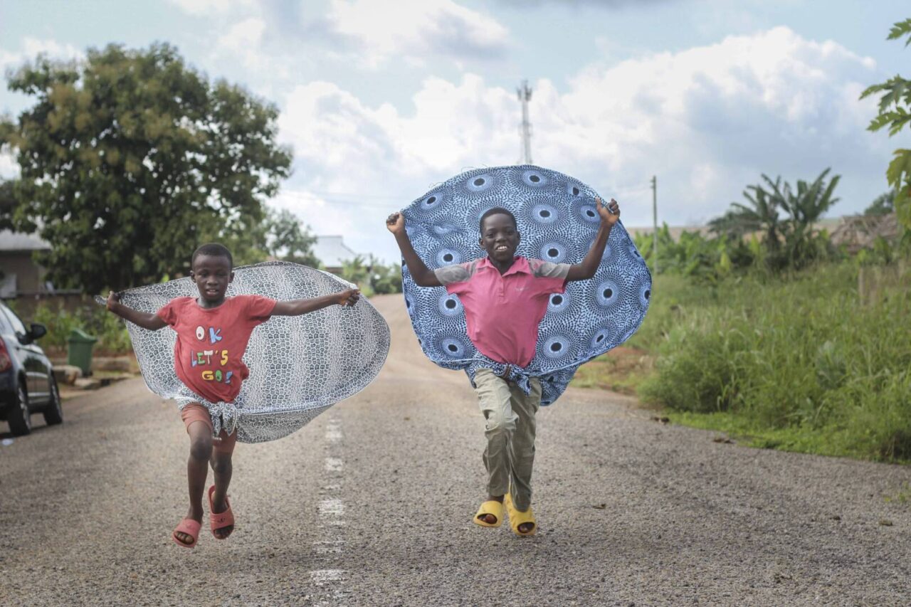 Ghanian boys in red shirts jump in the middle of the road with makeshift capes tied around their waists and extending behind them.