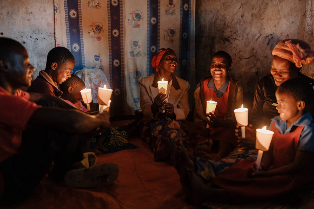 A family of seven or eight people, mostly children, sing happily while sitting on the floor of their simple cement home in Malawi. Each member holds a candle, the golden flames casting warm light upon their faces.