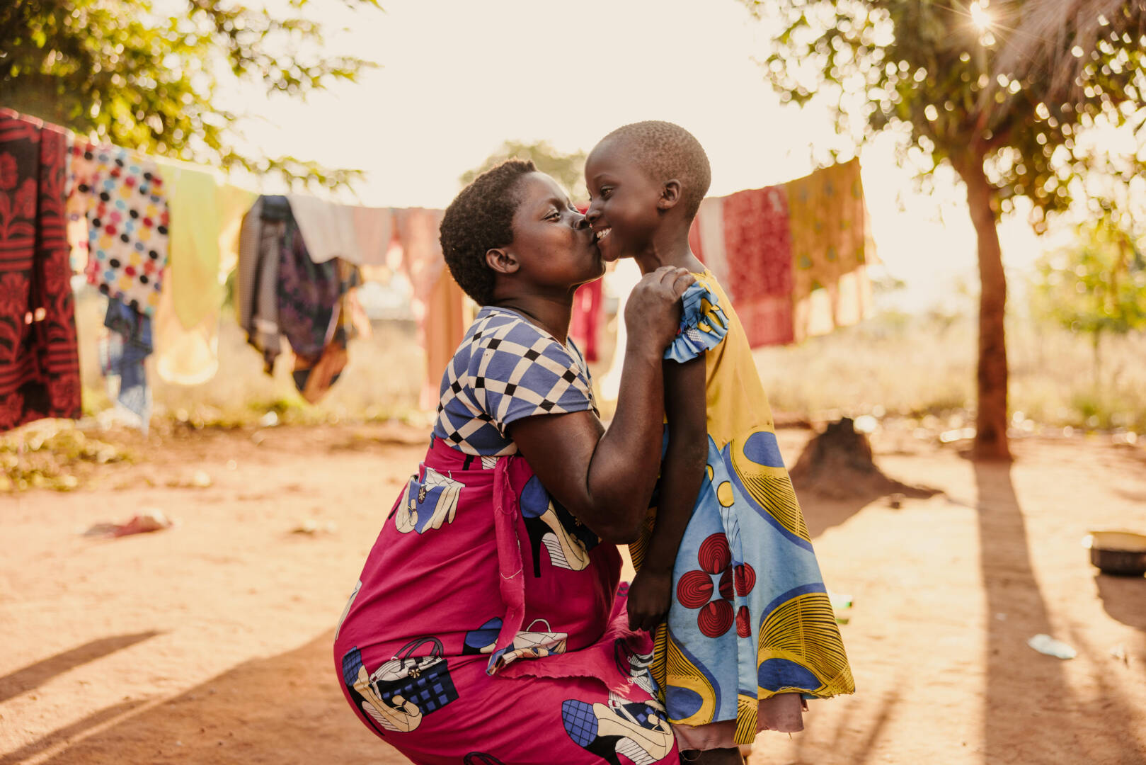 A woman squats to give a smiling young girl a loving, motherly kiss.