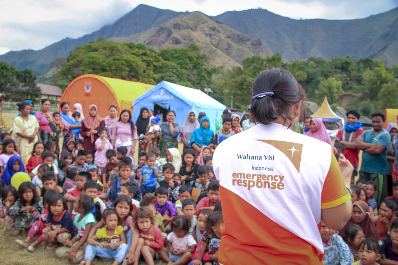 Children and women sit and stand outdoors, attentively listening to a World Vision staff member wearing an &ldquo;Indonesia emergency response&rdquo; shirt.