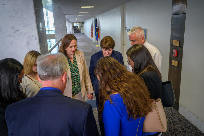Six women and two men in business attire join hands in a hallway with flags in the background.