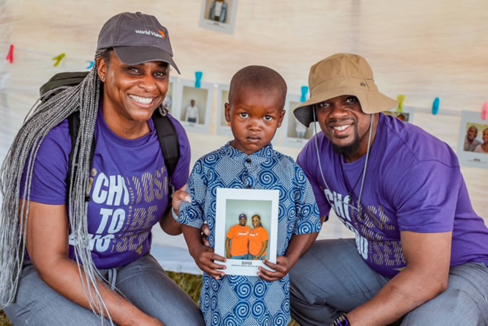 A boy stands between a man and woman who squat beside him. The boy holds a photo in his hands of the same man and woman.