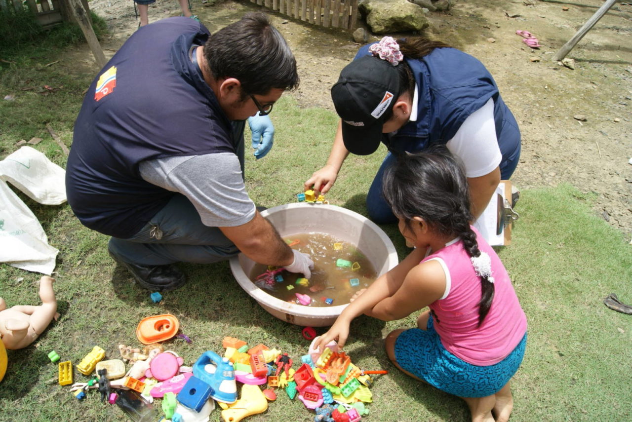 An aerial view of two adults in navy shirts and a young girl with a braid wearing pink, all leaning over a bin filled with water and colorful plastic toys.