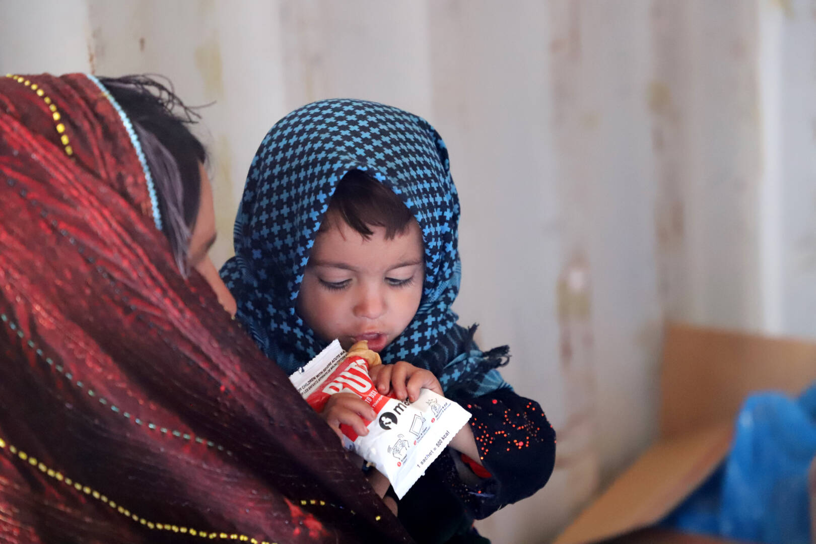 A child in her mother&rsquo;s arms eats a packet of food. They both wear colorful headscarves. 