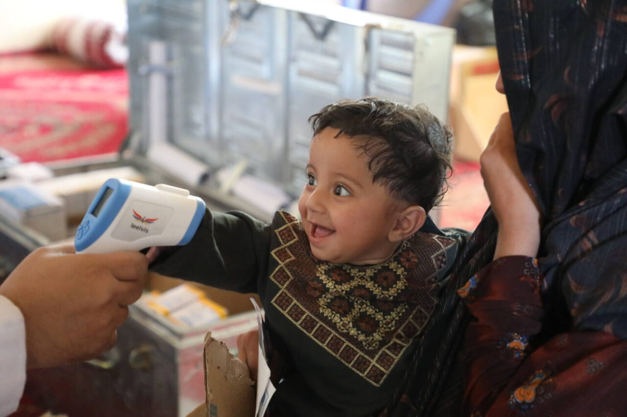 A health worker checks a smiling young Afghan girl’s temperature.