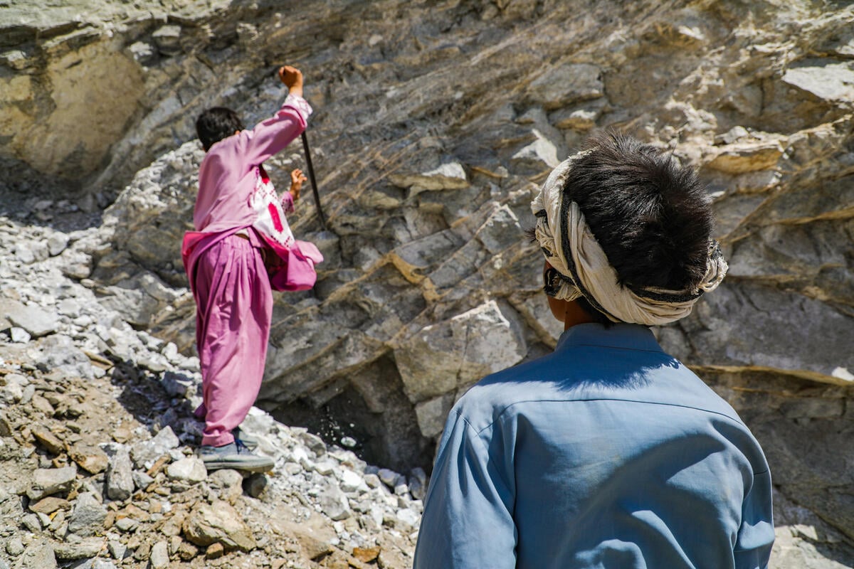 Two boys are working in a rocky area. One boy in dark pink digs with a shovel while the other in blue watches. Both boys face away from the camera.