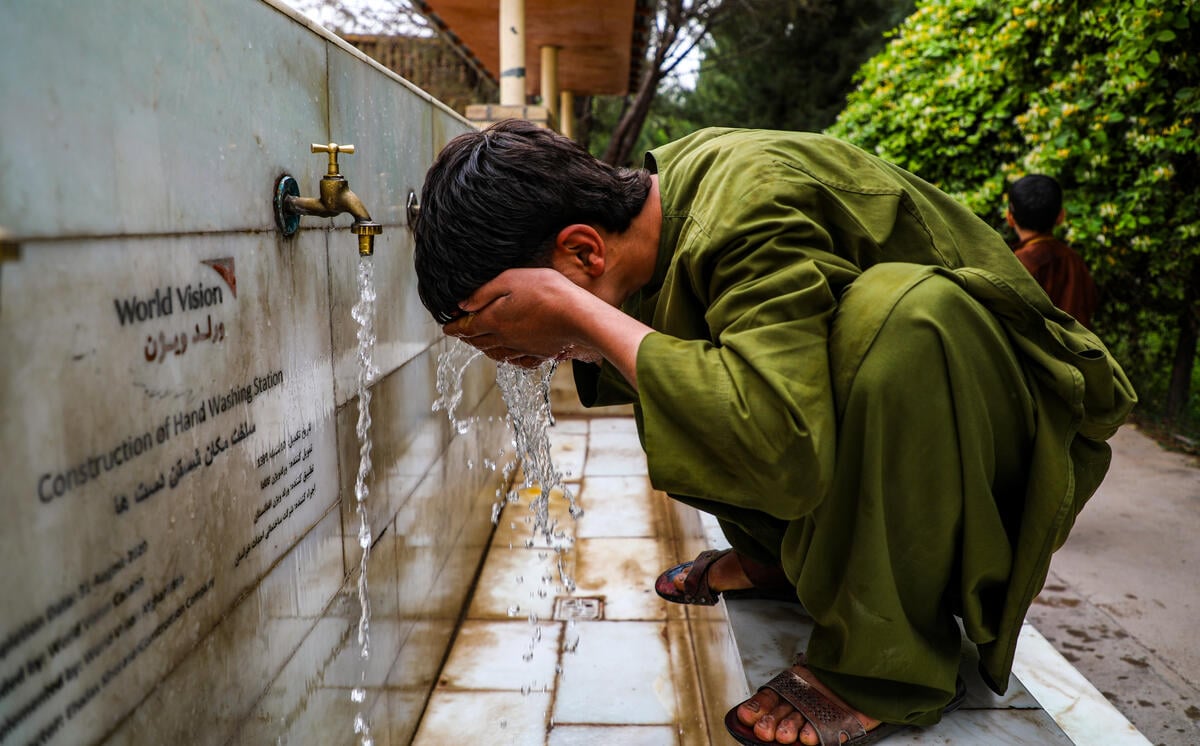 A boy in green splashes water on his face from a faucet at a water source, with a World Vision logo visible on the concrete wall in front of him.