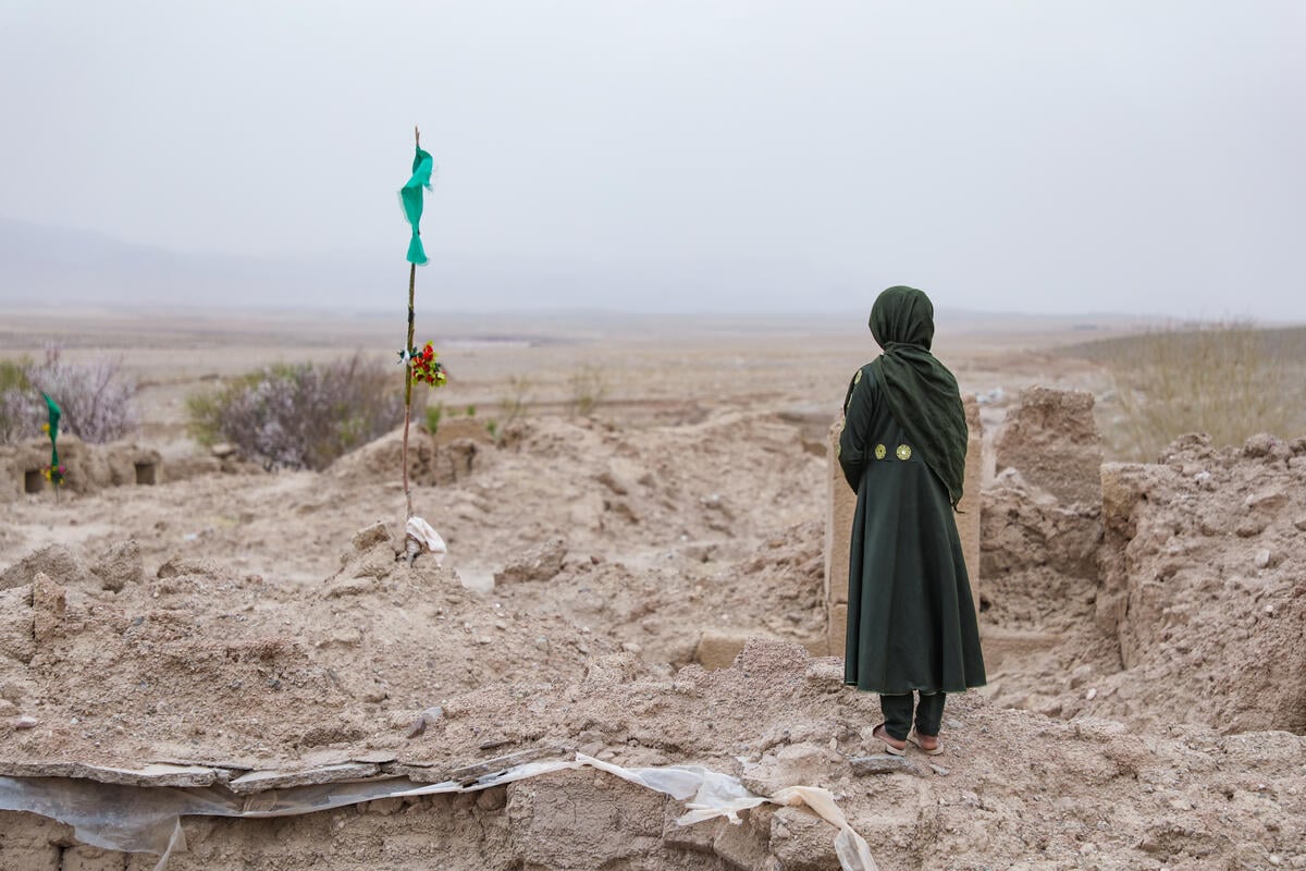 A girl in a green veil and dress stands with her back to the camera looking at a memorial, a pole adorned with fabric and flowers.