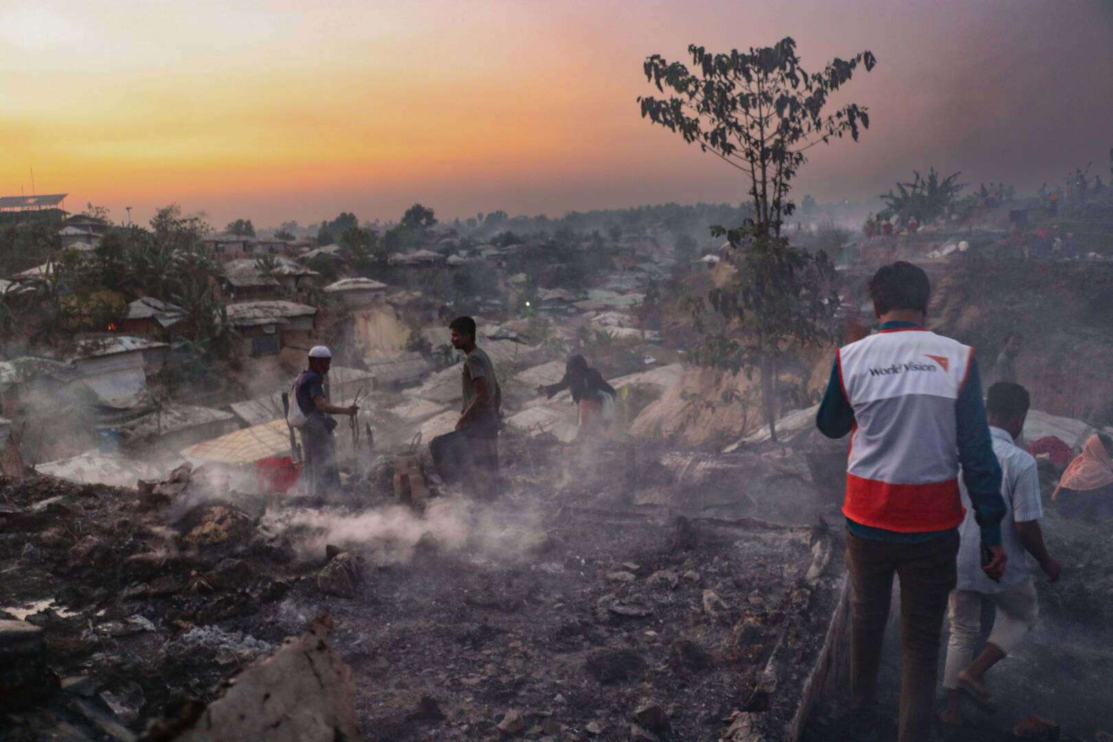 A man in a World Vision vest walks toward others in smoldering ruins under an orange dusk sky.