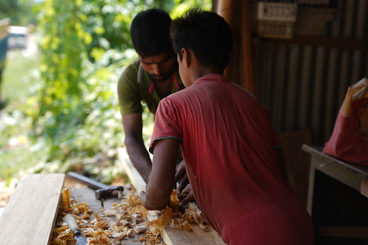 The back of a boy in a red shirt is turned as he works alongside a man, shaving wood at a woodworking factory.