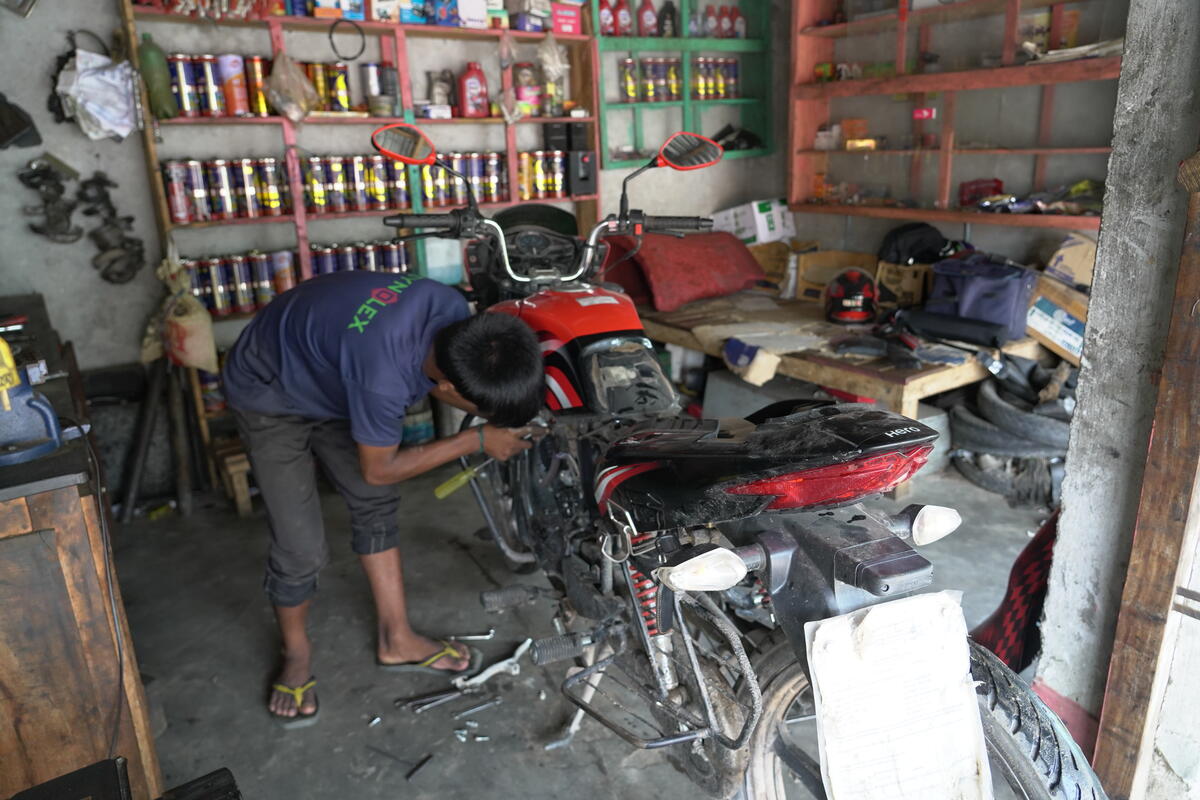 A boy wearing flip-flops leans over a motorcycle in a repair shop, where tools are scattered across the ground.